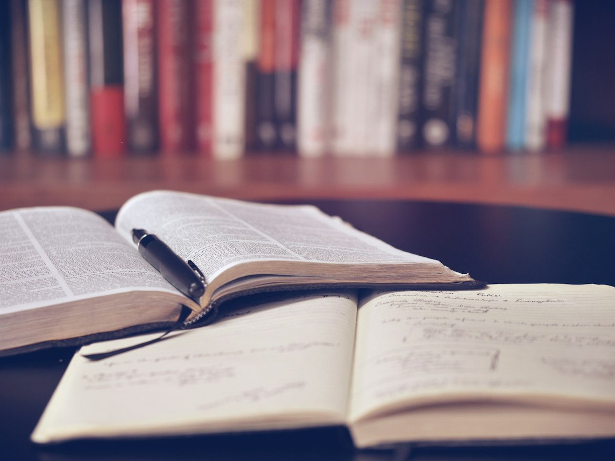 Livros e cadernos abertos sobre uma mesa, com uma estante ao fundo.